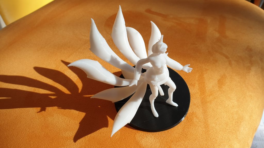 Eine weiße 3D-gedruckte Figur mit neun großen Schwänzen steht auf einem schwarzen runden Sockel und wirft einen Schatten auf eine orangefarbene Fläche. Die Figur scheint Tierohren und einen ausgestreckten Arm zu haben.