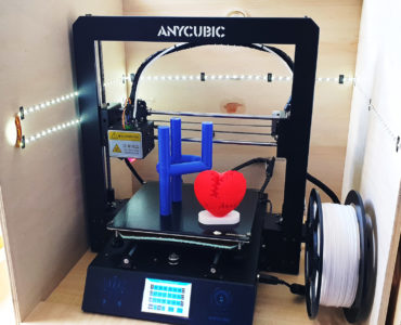 Ein 3D-Drucker mit der Aufschrift Anycubic in einem Holzgehäuse, der blaue und rote Kunststoffobjekte druckt, darunter ein Herz. Eine Spule mit weißem Filament ist auf der rechten Seite des Druckers angebracht.