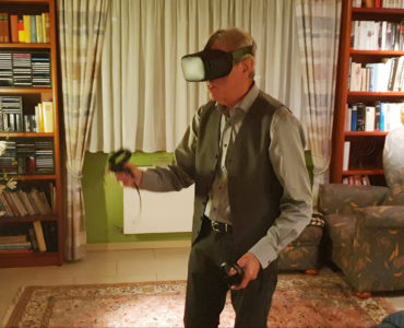 Ein älterer Mann, der ein VR-Headset trägt und Controller in der Hand hält, steht in einem gemütlichen Wohnzimmer mit Bücherregalen, einem geblümten Sessel und einem gemusterten Teppich und taucht in die 3D-Darstellung ein, während das Sonnenlicht durch das Fenster mit den Vorhängen hinter ihm fällt.