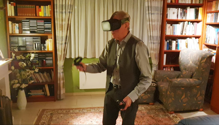 Ein älterer Mann, der ein VR-Headset trägt und Controller in der Hand hält, steht in einem gemütlichen Wohnzimmer mit Bücherregalen, einem geblümten Sessel und einem gemusterten Teppich und taucht in die 3D-Darstellung ein, während das Sonnenlicht durch das Fenster mit den Vorhängen hinter ihm fällt.