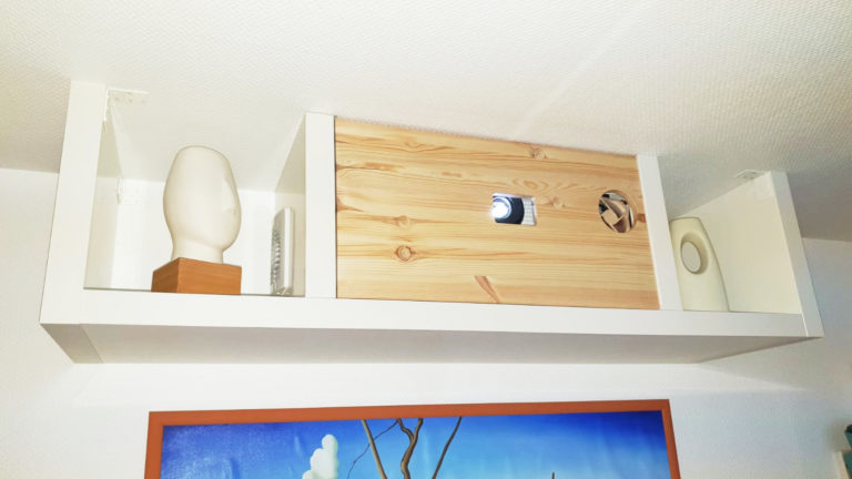 Ein weißes Wandregal zeigt eine 3D-Darstellung einer Kopfskulptur auf einem Holzsockel, ein kleines vertikales Objekt, ein zentrales Holzkästchen mit einem Loch, einen runden dekorativen Gegenstand und ein weißes ovales Stück. Darunter hängt ein Gemälde.