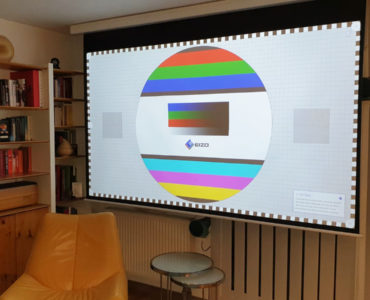 Ein Wohnzimmer mit einem gelben Sessel, Glastischen, Bücherregalen und einer großen Projektorleinwand, die ein buntes EIZO-Monitor-Testbild in einer realistischen 3D-Darstellung zeigt.