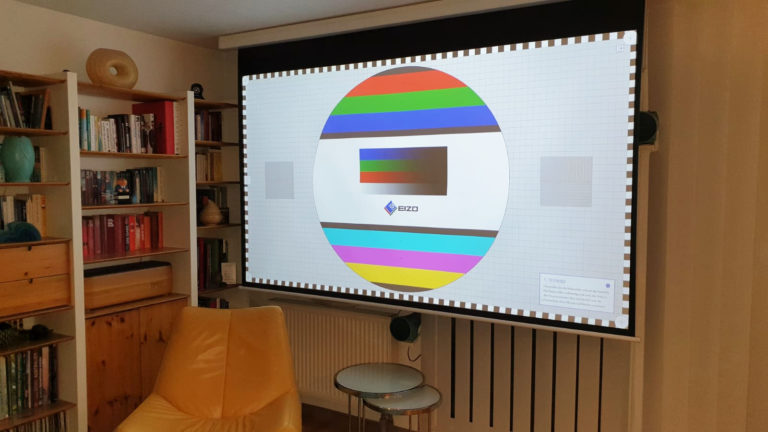 Ein Wohnzimmer mit einem gelben Sessel, Glastischen, Bücherregalen und einer großen Projektorleinwand, die ein buntes EIZO-Monitor-Testbild in einer realistischen 3D-Darstellung zeigt.