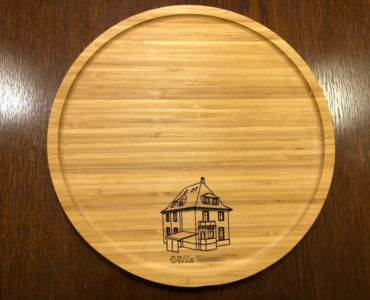 Ein rundes Holztablett mit der eingravierten Abbildung eines Hauses und dem Wort Villa am Boden, das auf einer dunklen Holzfläche steht.
