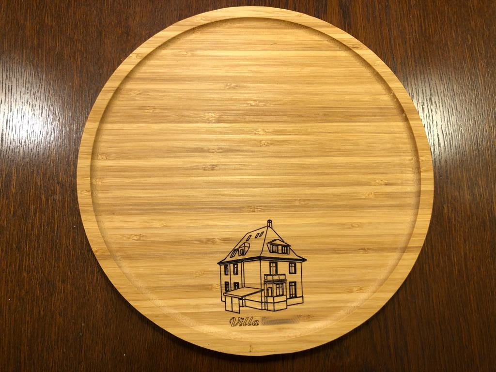 Ein rundes Holztablett mit der eingravierten Abbildung eines Hauses und dem Wort Villa am Boden, das auf einer dunklen Holzfläche steht.