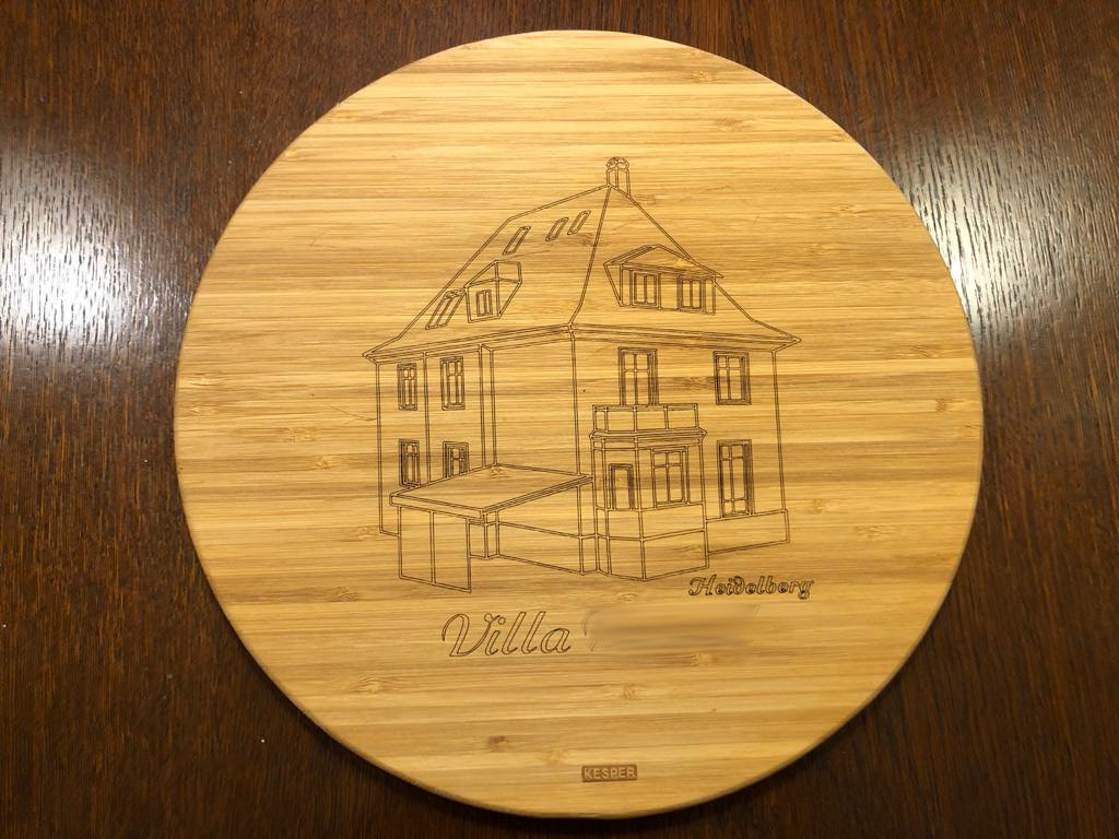 Ein rundes Holzbrett mit einer eingravierten Strichzeichnung eines Hauses mit der Bezeichnung Villa Heidelberg. Das Brett hat eine glatte, helle Holzoberfläche, und die Skizze zeigt architektonische Details der Villa.