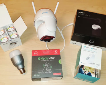 Mehrere Smart-Home-Geräte auf einer hellen Holzoberfläche, darunter eine Sicherheitskamera, ein intelligentes Schloss, eine LED-Glühbirne, ein Sechserpack Batterien, ein GPS-Tracker für Haustiere und ein intelligenter Stecker in den jeweiligen Boxen.