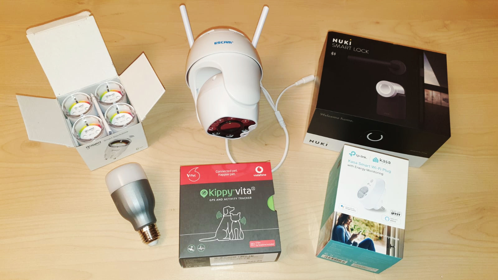 Mehrere Smart-Home-Geräte auf einer hellen Holzoberfläche, darunter eine Sicherheitskamera, ein intelligentes Schloss, eine LED-Glühbirne, ein Sechserpack Batterien, ein GPS-Tracker für Haustiere und ein intelligenter Stecker in den jeweiligen Boxen.