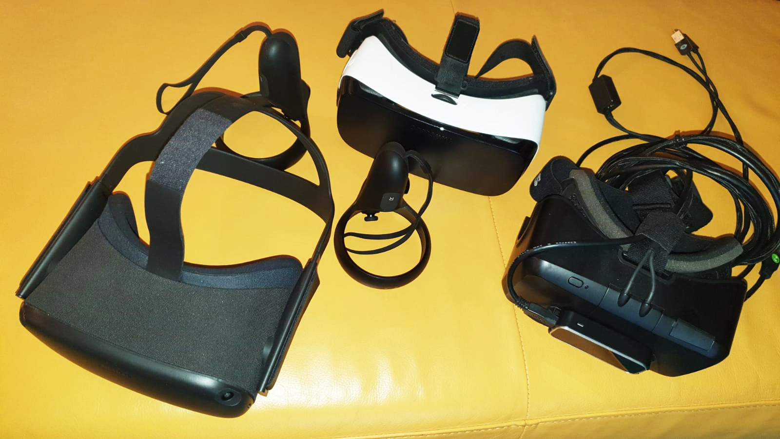 Drei Virtual-Reality-Headsets und zwei Motion-Controller werden auf einer gelben Fläche präsentiert, wobei die Kabel neben einem Headset für eine beeindruckende 3D-Darstellung sichtbar sind.