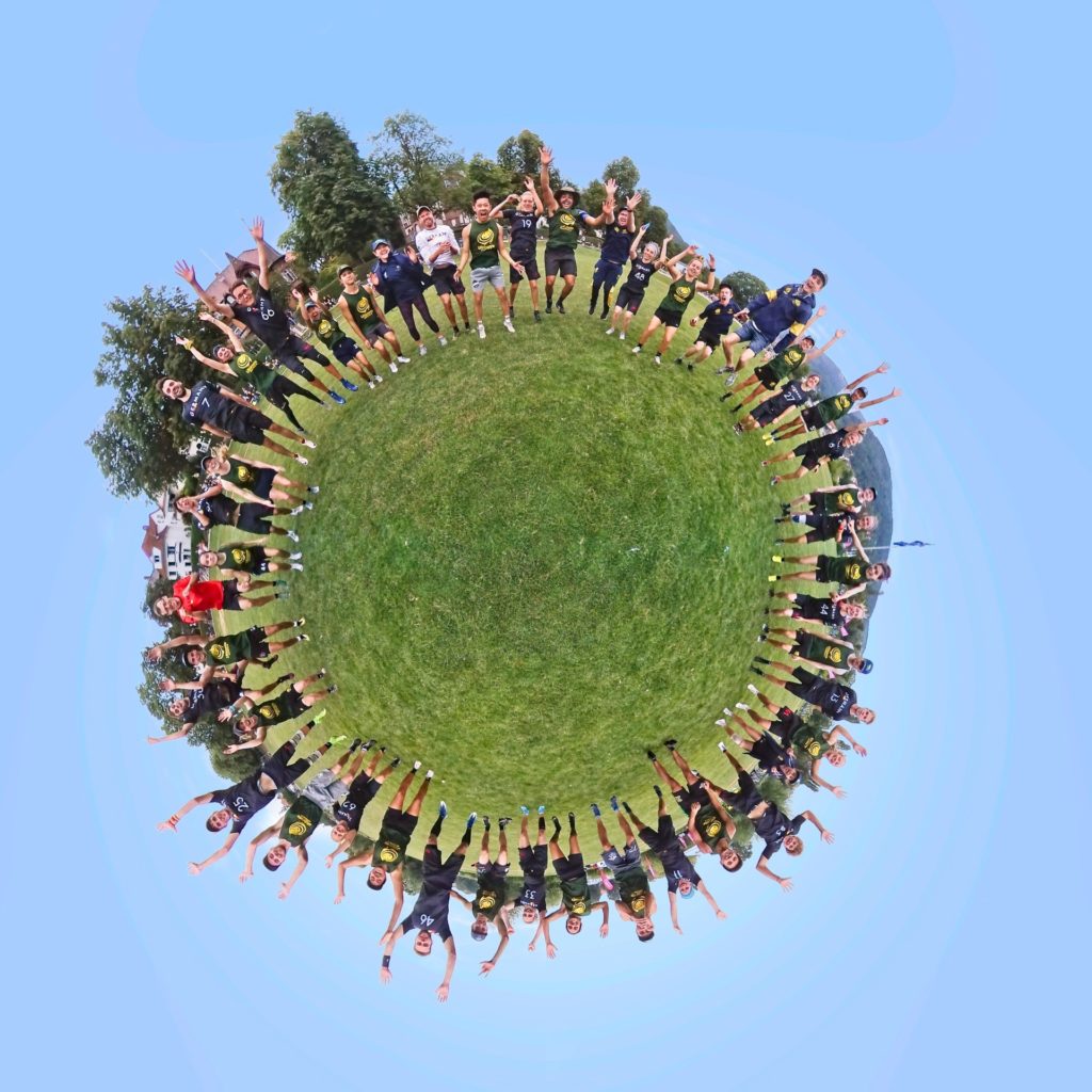 Eine Gruppe von Menschen in Sportuniformen steht in einer kreisförmigen Formation auf einer Wiese und feiert mit erhobenen Armen unter einem klaren blauen Himmel; die Szene fängt den Effekt einer 360° Aufnahme eines kleinen Planeten ein.