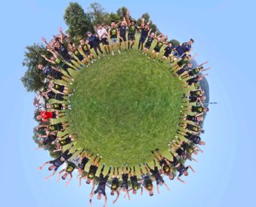 Eine Gruppe von Menschen in Sportuniformen steht in einer kreisförmigen Formation auf einer Wiese und feiert mit erhobenen Armen unter einem klaren blauen Himmel; die Szene fängt den Effekt einer 360° Aufnahme eines kleinen Planeten ein.