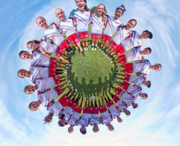 Eine Mädchenfußballmannschaft steht im Kreis auf dem Rasen, von oben gesehen mit einem 360°-Aufnahmeeffekt, der ein kreisförmiges Muster vor einem blauen Himmel erzeugt. Die Spielerinnen lächeln und tragen passende weiße und rote Trikots mit sichtbaren Nummern.