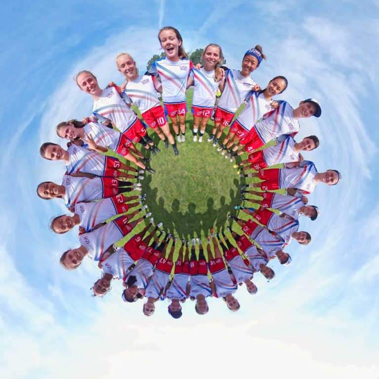 Eine Mädchenfußballmannschaft steht im Kreis auf dem Rasen, von oben gesehen mit einem 360°-Aufnahmeeffekt, der ein kreisförmiges Muster vor einem blauen Himmel erzeugt. Die Spielerinnen lächeln und tragen passende weiße und rote Trikots mit sichtbaren Nummern.