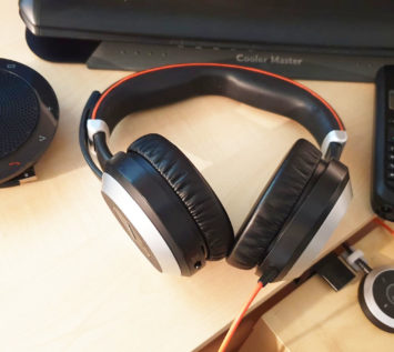 Ein schwarz-silberner Over-Ear-Kopfhörer liegt auf einem hellen Holztisch, umgeben von Geräten, die für die Online-Kommunikation unerlässlich sind, darunter eine Freisprecheinrichtung, ein schnurloses Telefon, ein Computermonitor und ein Audiosteuergerät.