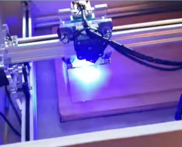 Eine Nahaufnahme einer Lasergravurmaschine in Betrieb, die ein blaues Licht ausstrahlt, während sie eine rechteckige Holzoberfläche graviert.
