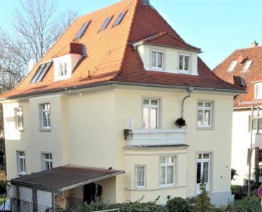 Ein dreistöckiges, cremefarbenes Haus mit rotem Ziegeldach, mehreren Fenstern, einem Balkon und einem kleinen überdachten Carport, umgeben von einem Zaun und Grünflächen - die perfekte Inspiration für 2D-Design- oder LaserCut-Projekte.