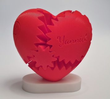 Ein rotes 3D-gedrucktes Herz mit ineinander greifenden Zahnrädern, eines davon mit dem Namen Yannick graviert, auf einem weißen ovalen Sockel vor einem schlichten Hintergrund.