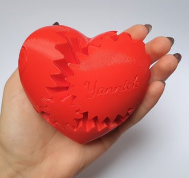 Eine Hand hält ein rotes, herzförmiges 3D-gedrucktes Objekt mit sichtbaren, ineinander greifenden Zahnrädern und dem eingravierten Namen Yannick auf seiner Oberfläche.