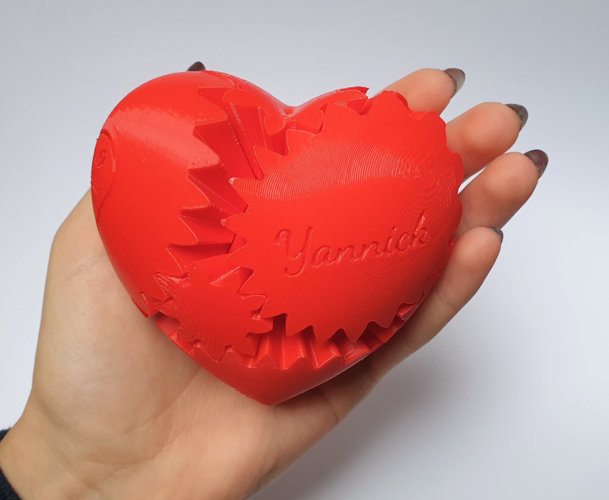 Eine Hand hält ein rotes, herzförmiges 3D-gedrucktes Objekt mit sichtbaren, ineinander greifenden Zahnrädern und dem eingravierten Namen Yannick auf seiner Oberfläche.