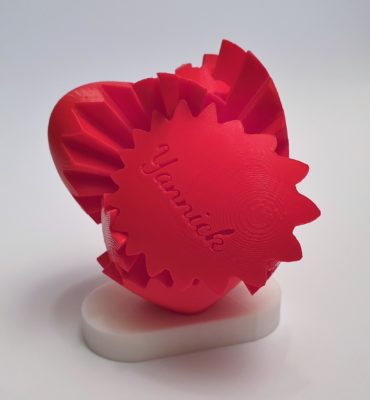 Ein rotes 3D-gedrucktes mechanisches Herz mit sichtbaren Zahnrädern und dem eingravierten Namen Yannick auf einem Zahnrad, montiert auf einem weißen Ständer vor einem schlichten hellen Hintergrund.