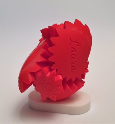 Eine rote 3D-gedruckte herzförmige Zahnradskulptur mit dem Namen Laura auf einem Zahnrad, montiert auf einem weißen Sockel vor einem schlichten Hintergrund.