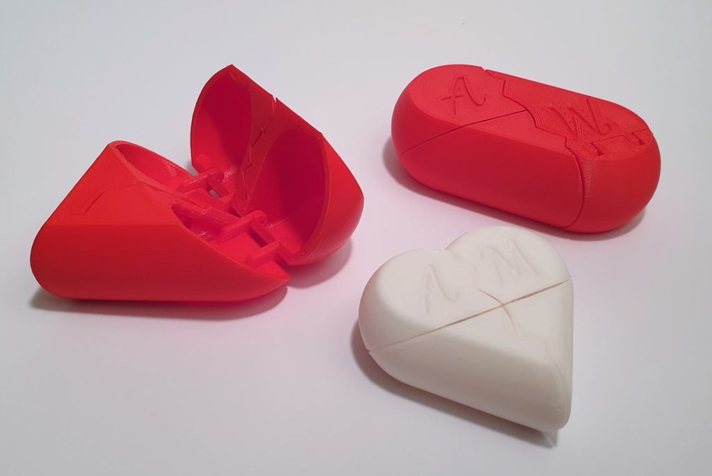 Drei 3D-gedruckte herzförmige Puzzleschachteln, eine rote und offene, eine rote und geschlossene und eine weiße und geschlossene, jeweils mit den Buchstaben A und M auf der Oberseite, alle auf weißem Hintergrund.