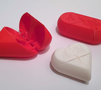 Drei 3D-gedruckte herzförmige Puzzleschachteln, eine rote und offene, eine rote und geschlossene und eine weiße und geschlossene, jeweils mit den Buchstaben A und M auf der Oberseite, alle auf weißem Hintergrund.