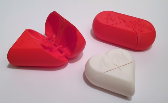 Drei 3D-gedruckte herzförmige Puzzleschachteln, eine rote und offene, eine rote und geschlossene und eine weiße und geschlossene, jeweils mit den Buchstaben A und M auf der Oberseite, alle auf weißem Hintergrund.