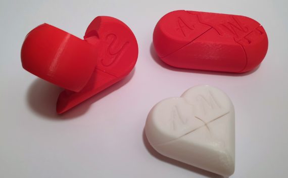 Drei 3D-gedruckte Puzzleteile: eine zweigeteilte rote Kapsel mit Initialen, ein zweigeteiltes rotes herzförmiges Teil mit einer Initiale und ein zweigeteiltes weißes herzförmiges Teil mit den Initialen AM.