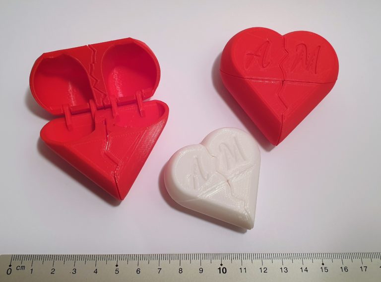 Drei 3D-gedruckte herzförmige Behälter, zwei rote und ein weißer, mit AM auf der Oberseite und einem Rissmuster in der Mitte, werden gezeigt. Ein rotes Herz ist geöffnet und enthüllt ein Scharnier. Zur Veranschaulichung ist am unteren Rand ein Lineal angebracht.