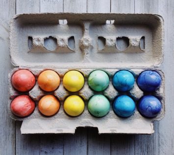 Ein Dutzend in Regenbogenfarben gefärbte Eier, die in einem Pappkarton auf einer rustikalen Holzfläche angeordnet sind. Auf dem leuchtenden Display steht "Frohe Ostern", wobei die Farben von Rot und Orange auf der linken Seite zu Blau und Lila auf der rechten Seite übergehen.