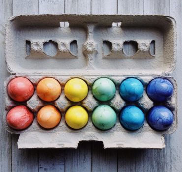 Ein Dutzend in Regenbogenfarben gefärbte Eier, die in einem Pappkarton auf einer rustikalen Holzfläche angeordnet sind. Auf dem leuchtenden Display steht "Frohe Ostern", wobei die Farben von Rot und Orange auf der linken Seite zu Blau und Lila auf der rechten Seite übergehen.