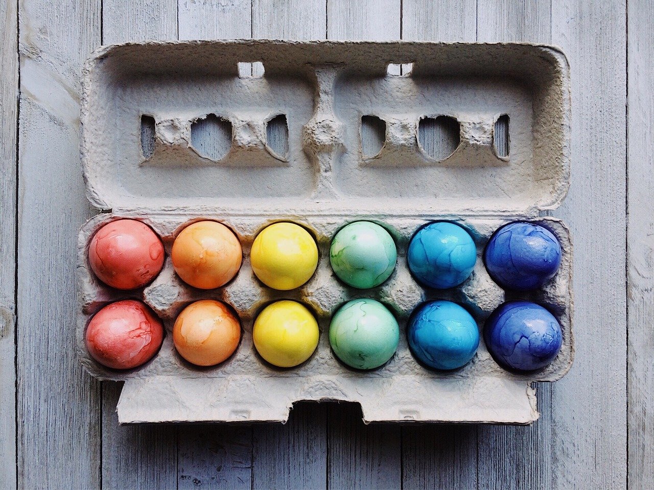 Ein Dutzend in Regenbogenfarben gefärbte Eier, die in einem Pappkarton auf einer rustikalen Holzfläche angeordnet sind. Auf dem leuchtenden Display steht "Frohe Ostern", wobei die Farben von Rot und Orange auf der linken Seite zu Blau und Lila auf der rechten Seite übergehen.