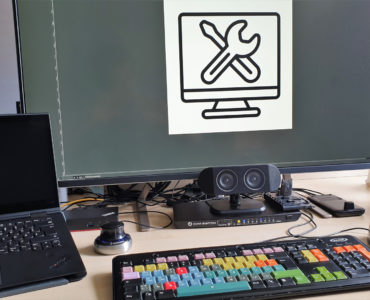 Ein Schreibtisch mit einer bunten Tastatur, einer Maus, zwei Laptops, einem externen Lautsprecher und einem großen Monitor, der einen Computerbildschirm mit einem Schraubenschlüssel- und Schraubenziehersymbol zeigt. Kabel und technische Geräte sind auf dem Schreibtisch zu sehen.