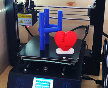 Ein 3D-Drucker mit digitaler Anzeige zeigt zwei fertige Drucke: eine blaue geometrische Form, die einer Hand ähnelt, die den Felsen auf Geste macht, und ein rotes Herz mit ineinandergreifenden puzzleartigen Teilen.