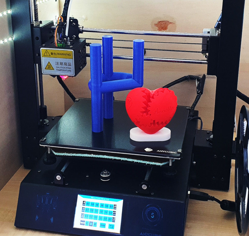 Ein 3D-Drucker mit digitaler Anzeige zeigt zwei fertige Drucke: eine blaue geometrische Form, die einer Hand ähnelt, die den Felsen auf Geste macht, und ein rotes Herz mit ineinandergreifenden puzzleartigen Teilen.